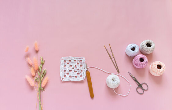 Top view of crocheting process of white square lace doily on pink pastel background. Skeins of yarn for knitting, crochet hooks and bouquet of ears of oats and lagurus nearby. Copy space, flat lay
