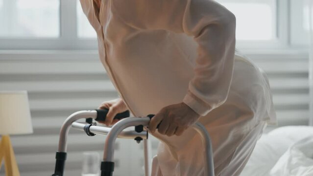 Lonely elderly woman stands up from bed and navigates bedroom using a walker