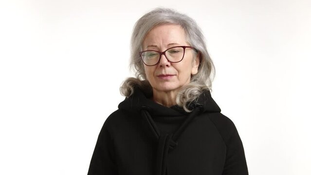 A thoughtful senior woman wearing glasses, captured with a serious and reflective expression, set against a plain white background. High quality 4k footage