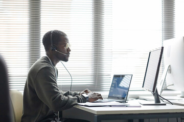 Side view portrait of Black young man wearing headset and using computers as client support...