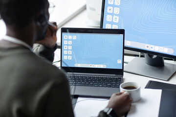 Over shoulder view of young Black man using laptop with blue desktop screen in minimal office setting and drinking coffee copy space