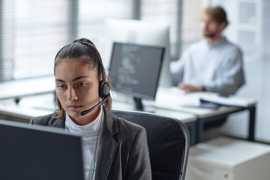 Portrait of young woman wearing headset with microphone and using computer while working in customer support office copy space