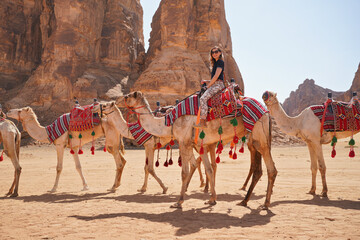 Group of camels, seats ready for tourists, walking in AlUla desert on a sunny day afternoon, young woman riding one animal, sandstone rocks formation background - Powered by Adobe