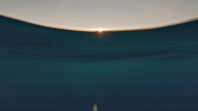 Swimming at sea during sunset, sun setting down behind hills in distance, half camera underwater, dark water visible - thalassophobia concept