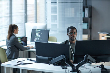 Portrait of Black young man as IT programmer using multiple computers at workplace in office and...