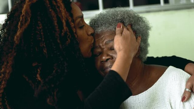 Tender loving moment of African American adult daughter kissing elderly 80s senior mother in the forehead and posing close-up faces smiling at camera
