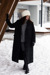 beautiful young fashionable woman in white fur hat and dark winter coat poses on the streets of the city in winter