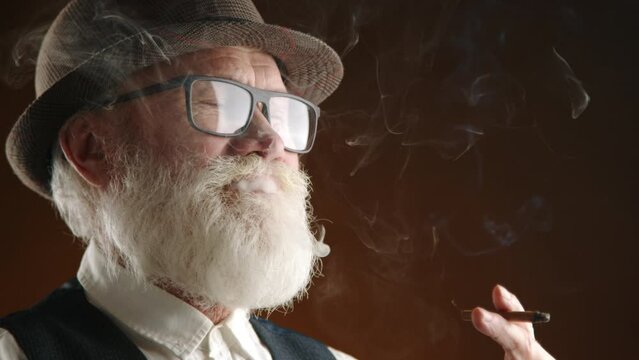 An elegant senior man with a full white beard and spectacles exhales smoke while enjoying a cigar, wearing a fedora hat and a vintage vest, conveying a mood of contemplation and relaxation. Camera 8K.