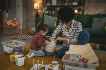 Mother and toddler daughter at home collect toys put in box clean room