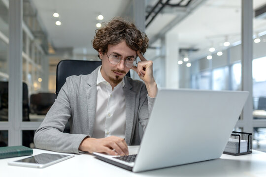 Serious young male professional with curly hair concentrates on his work at a laptop in a well-lit office setting.