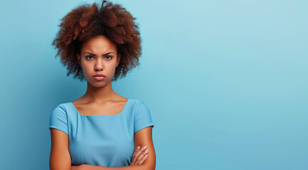 woman with curly hair looks seriously at the camera, arms crossed, standing against a soft blue background - obrazy, fototapety, plakaty