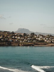 A surfer surfs a wave on the coast with the houses, the mountain and the sunny sky in the background.
