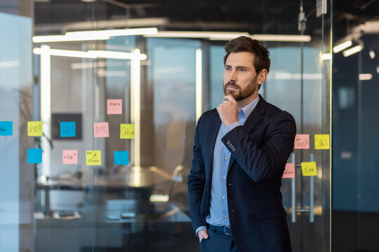 A contemplative businessman in a suit stands pondering before a glass wall filled with sticky notes and data charts.