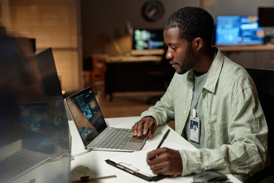 Side view portrait of Black young man using computer and taking notes while working late in dark office, copy space