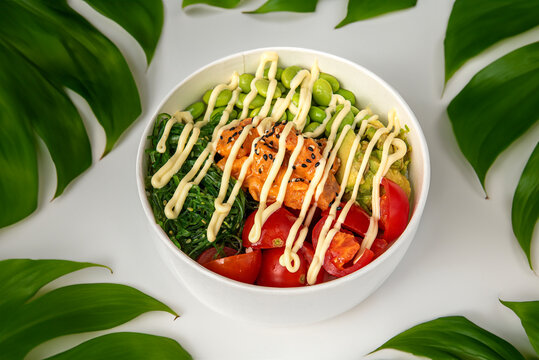 Poke bowl made with venus rice seasoned with soy sauce, seared tuna, red onion , purple cabbage, wakame seaweed and mango isolated on white table with tropical leaves - Powered by Adobe