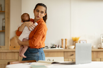 Mother freelancer holding in arms crying infant daughter and looking at laptop, working remotely...