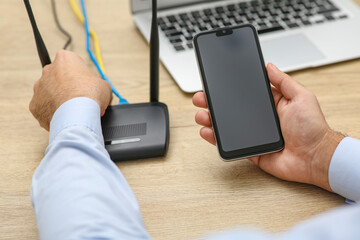 Man with smartphone and laptop connecting to internet via Wi-Fi router at wooden table, closeup