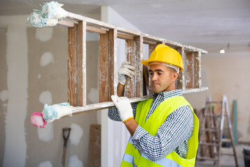 Man repairer in vest and hardhat carrying stepladder. Male builder working in construction site.