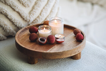 Burning aromatic candle with the sweet fresh scent of exotic tropical fruits on wooden bamboo tray. Home aromatherapy, detention, relaxation, meditation. Concept of wellness, wellbeing, cozy interior