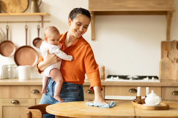 Spring cleaning. Mother holding little baby daughter and cleaning table in kitchen interior, mom...