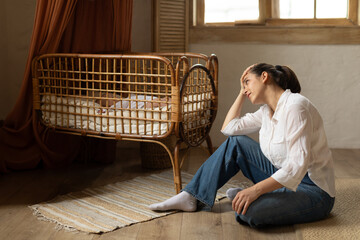 Unhappy young mother sitting on floor near child bed while baby sleeping, woman suffering postnatal...