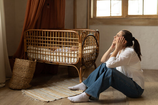 Young mother suffering postnatal depression symptoms, woman sitting on floor near child bed while baby sleeping, free space