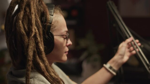 Waist up of young Caucasian woman with dreadlocks putting on headphones and checking mic while preparing for audio podcast recording with Biracial male colleague in dark studio at nighttime