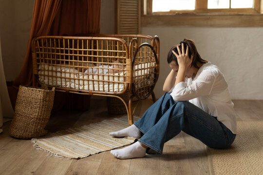 Young mother feeling desperate, sitting on floor next to baby napping in bed, woman crying, suffering postnatal depression, copy space