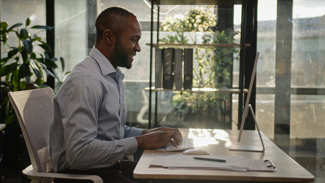 Middle-aged mature African American ethnic man worker CEO director sit at sunny office business agency company typing email on laptop computer search data online networking businessman smiling smile