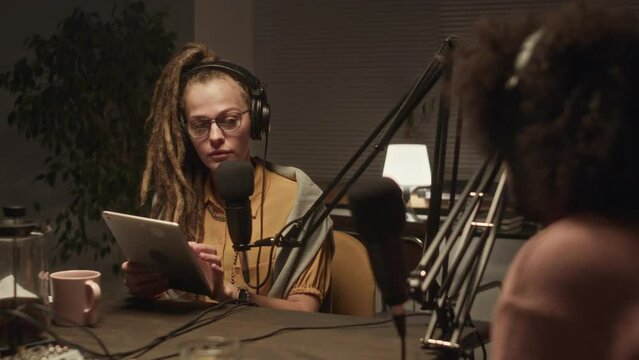 Over shoulder waist up of young Caucasian woman with dreadlocks looking at notes on digital tablet while interviewing male guest during audio podcast late in dark studio with dimmed light