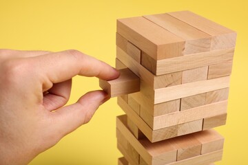 Playing Jenga. Man removing wooden block from tower on yellow background, closeup