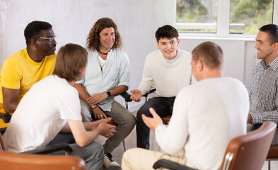 Cheerful carefree male colleagues of various ages and nationalities friendly talking sitting in circle on chairs in office lounge during break, sharing personal stories..