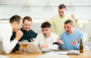 Group of men watching sports match online on laptop during friendly meeting with beer at home, excitedly celebrating winning score in game ..