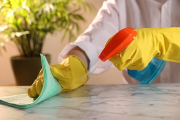 Woman with spray bottle and microfiber cloth cleaning white marble table at home, closeup