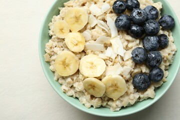 Tasty oatmeal with banana, blueberries, coconut flakes and honey served in bowl on beige table, top view