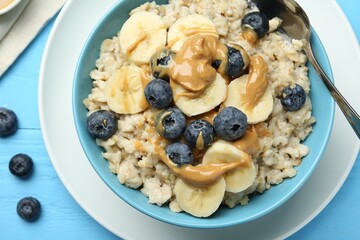 Tasty oatmeal with banana, blueberries and peanut butter served in bowl on light blue wooden table,...