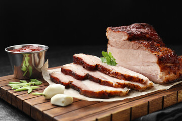Pieces of baked pork belly served with sauce and parsley on black textured table, closeup