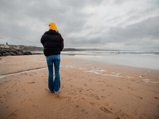 Teenager girl in black jacket and yellow hat walking on a sandy beach, cloudy sky. Lahinch, county...