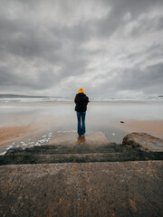 Teenager girl in black jacket standing on a footpath to a vast sandy beach and ocean, dramatic sky...