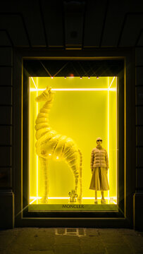 Florence, Italy - August 29 2023 : A stylish window display for luxury brand moncler with giraffe in puffer coat and manikin in coat, glasses and skirt at night. Bright neon yellow and white light