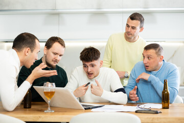 Group of men gathered for friendly meeting with beer at cozy home kitchen, trying to work through challenging problem, viewing laptop screen with concerned expressions and gestures ..