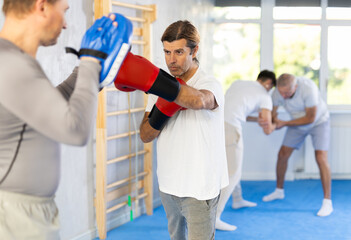 Fototapeta na wymiar Man in boxing training practices punches with man in mitts in sport club