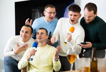 Group of cheerful male friends having fun at alcoholic bachelor party in cozy living room, drinking...