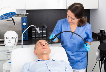 Confident female beautician performing face ultrasound lifting procedure for young man in clinic