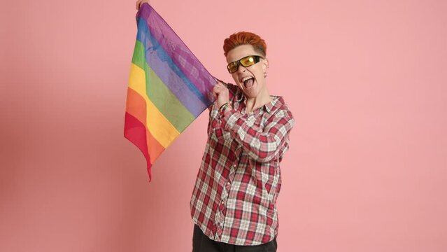 A confident mature lesbian with a stylish mohawk haircut proudly waves the LGBT pride flag, showcasing her vibrant spirit against a cheerful pink backdrop. Camera 8K RAW. High quality 4k footage