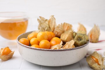 Ripe physalis fruits with calyxes in bowl on white tiled table, closeup