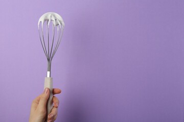 Woman holding whisk with whipped cream on violet background, closeup. Space for text