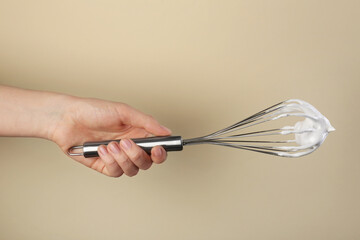 Woman holding whisk with whipped cream on beige background, closeup