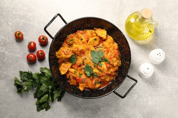 Delicious chicken curry in frying pan, parsley, tomatoes and oil on light grey table, flat lay