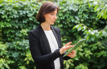 Fototapeta premium business professional woman against the backdrop of green nature. business woman with a tablet thinking, studying, counting, analyzing, looking.